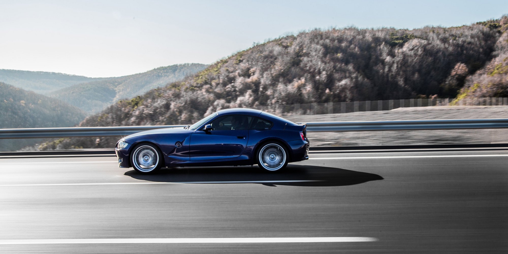 Blauer Sportwagen fährt bei Sonnenschein über eine leere Autobahn in bergiger Landschaft.